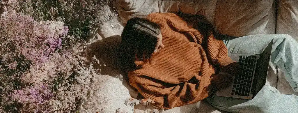 Person in orange sweater working on laptop surrounded by flowers